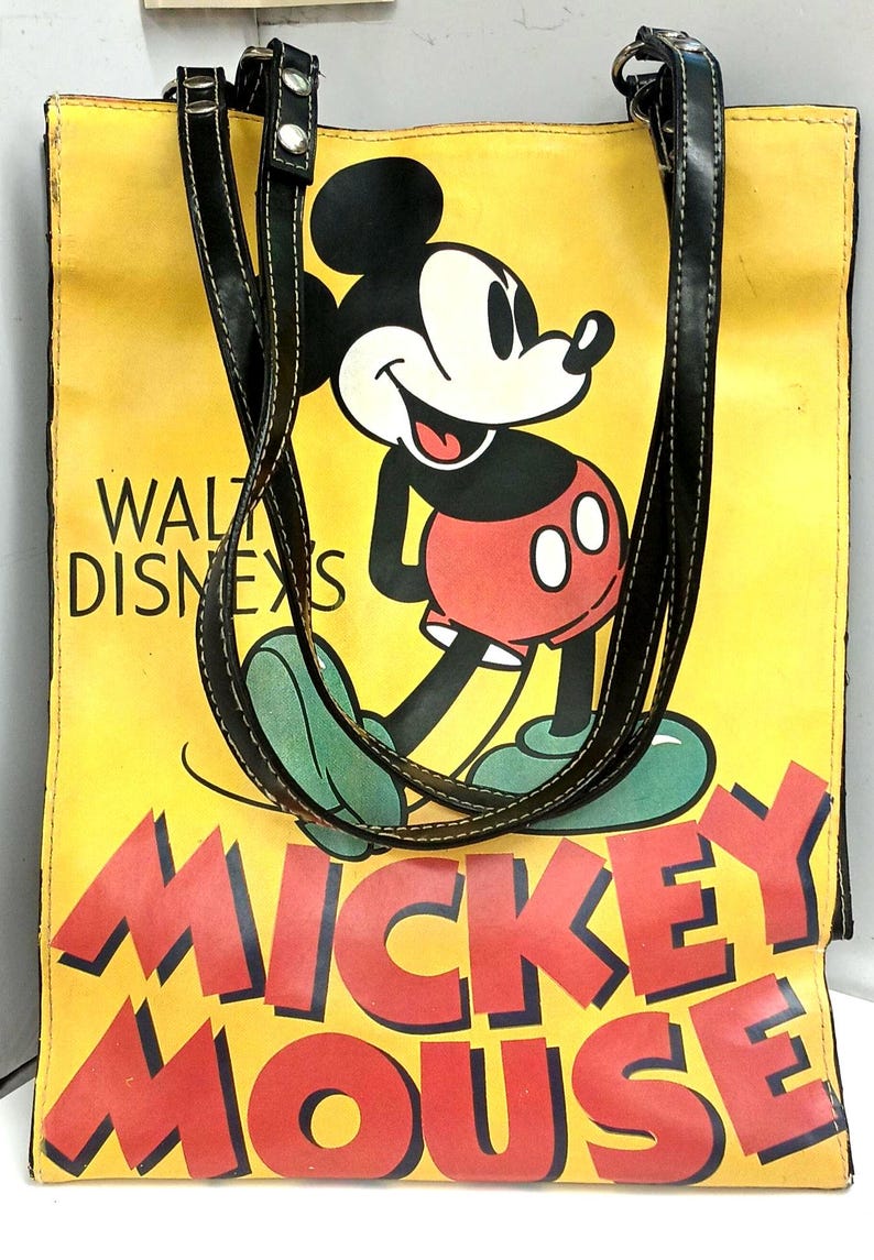 May include: Yellow tote bag featuring Mickey Mouse with black handles. The bag has the words "Walt Disney's" and "Mickey Mouse" in red lettering. Mickey Mouse is wearing red shorts, green shoes, and white gloves.