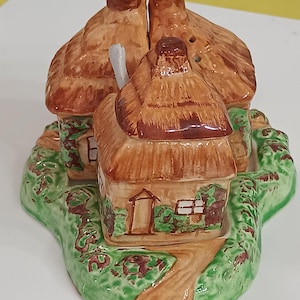 May include: A ceramic cottage-shaped salt and pepper shaker set. The set features three miniature houses with brown thatched roofs and green bases with a path and shrubbery. The houses have small windows and doors.