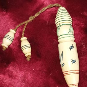 May include: Ivory-colored, hand-carved pendant with a decorative design. The pendant has a bullet-like shape with green and blue accents, and two smaller, matching beads attached by a brown cord. The design includes horizontal lines and cross-like symbols.