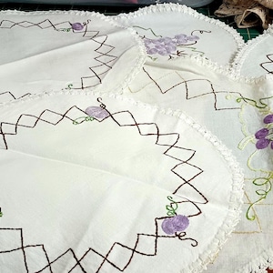 May include: A collection of white circular fabric doilies featuring embroidered purple grape clusters and green vines. Each piece is bordered by a brown diamond-patterned stitch and a white crocheted edge. The fabric items are arranged in a group.