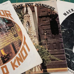 May include: Three vintage macrame pattern books. The covers feature the titles "To Knot-Or Not To Knot", "Macrame Pot Hangers", and "Macrame Portraits". The books are on a green cutting mat.