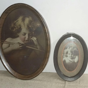 May include: Two antique oval picture frames. The larger frame has a dark wood finish and contains a sepia-toned image of a cherub holding a bow and arrow. The smaller frame is black with a sepia-toned image of a cherub.