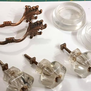 May include: An assortment of antique cabinet hardware, including two ornate bronze handles and three clear glass knobs. Also included are two clear plastic rings. The hardware is arranged on a white surface.