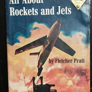May include: A vintage book cover featuring a colorful illustration of a rocket jet taking off against a cloudy sky. The title of the book is "All About Rockets and Jets" by Fletcher Pratt, illustrated by Jack Coggins. The book is part of the "Allabo Books" series, number 13.