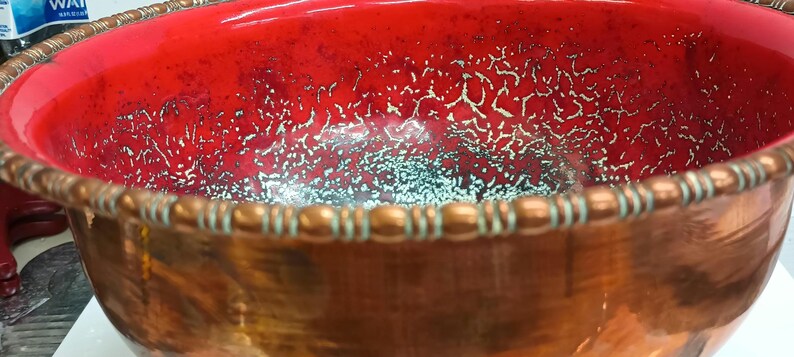 May include: A large copper bowl with a red interior and a decorative copper rim. The interior of the bowl has a speckled, metallic finish.