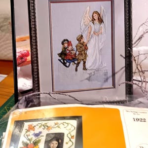May include: A cross-stitch kit titled "Guardian Angel Collection" featuring a framed image of an angel and children. Below, a kit for a picture frame with floral and bird embroidery is visible.