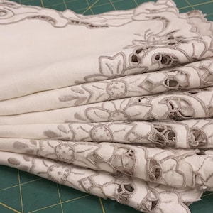 May include: A stack of folded white linen napkins with intricate gray floral embroidery along the edges. The napkins are neatly arranged, showcasing the delicate lace-like design. The background is a green cutting mat.