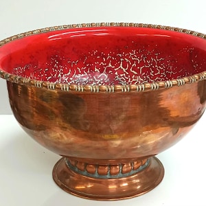 May include: A large copper punch bowl with a red interior and a decorative rim. The bowl sits on a pedestal base.