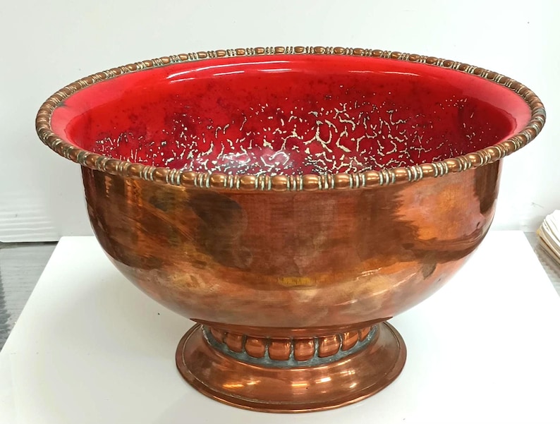 May include: A large copper punch bowl with a red interior and a decorative rim. The bowl sits on a pedestal base.