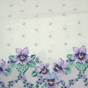 May include: White fabric with a repeating pattern of small purple flowers and a border of larger purple pansies with green leaves. The pansies have yellow centers. The fabric is likely used for home decor or sewing projects.
