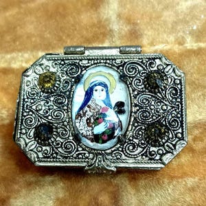 May include: A small, silver-toned metal box with a hinged lid. The box is decorated with an ornate design and has a clear glass window that reveals a painted image of a woman holding a flower. The box has a few small, clear rhinestones around the edges.