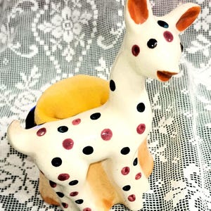 May include: A ceramic deer figurine featuring a cream-colored body with black and maroon polka dots, orange-tipped ears, and a yellow fabric cushion integrated into its back, designed to function as a decorative sewing pin holder.