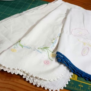 May include: A collection of white vintage handkerchiefs. One features a wide, ornate lace edge. Another has embroidered flowers in yellow, pink, and blue. The third has a blue crocheted border and embroidered flowers.