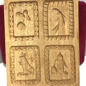 May include: Wooden cookie mold with four carved animal designs: a mushroom, a fish, a frog, and a person. The mold is rectangular with a decorative border around each animal. The wood has a natural, light brown color.