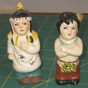 May include: Two vintage ceramic figurines depicting Native American figures. One figure wears a black hat with yellow accents and a white shirt with floral details. The other figure has black hair, a turquoise necklace, and a red skirt.