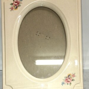May include: A white ceramic picture frame with an oval opening and floral designs in pink, blue, and yellow.