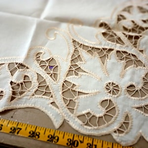 May include: Ivory-colored lace doily with a floral design. The doily features intricate cutwork and a delicate, scalloped edge. A yellow measuring tape is placed near the doily, with measurements in inches.