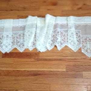 May include: A white lace valance curtain featuring a sheer, fine-mesh fabric body and a decorative, repeating geometric pattern along the bottom edge. The curtain has a straight top header and a series of pointed, lace-trimmed panels.