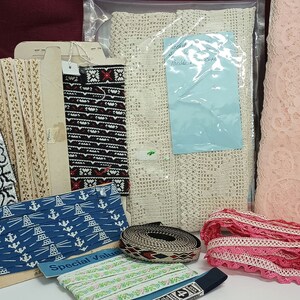 May include: A collection of various ribbons and lace in different colors and patterns. Includes a roll of pink lace, blue and white patterned ribbon, and beige lace. Some items are packaged, and others are on cardboard spools.