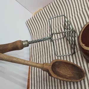 May include: A collection of vintage kitchen tools, including a wooden spoon, a potato masher, and a brown ceramic bowl. The spoon and masher have wooden handles. The bowl is dark brown with a lighter rim. The items rest on a striped cloth.