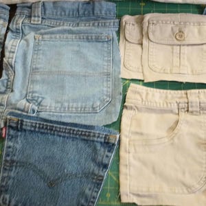 May include: Assortment of denim fabric scraps in various shades of blue, beige, and white. The fabric pieces are cut into different shapes and sizes, including squares, rectangles, and strips. Some pieces have pockets and seams.