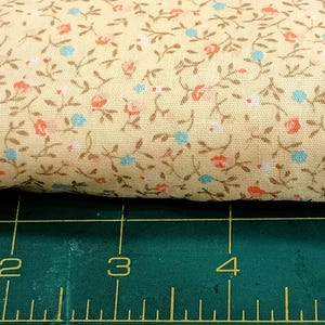 May include: A folded piece of fabric with a floral pattern. The fabric is a light yellow color with a repeating pattern of small red, blue, and brown flowers and leaves. The fabric is resting on a green cutting mat with inch measurements.
