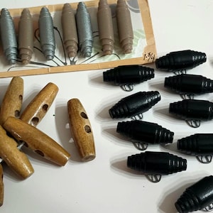 May include: A collection of various buttons in different styles and colors. There are wooden toggle buttons, black barrel buttons, and brown and silver-toned buttons. Some buttons are on a card with the measurement 1-3/4.