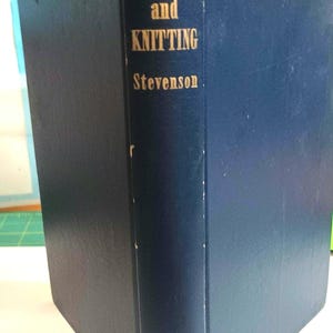 May include: A vintage navy blue book titled "CROCHET and KNITTING" by Stevenson. The book's spine features gold lettering and the publisher's name, "PEOPLES," at the bottom. The book's cover shows signs of wear.