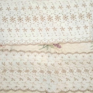 May include: Cream-colored floral embroidered fabric with scalloped edges. The fabric features a repeating pattern of small flowers and delicate scrollwork. The design includes a floral embroidered section with a lavender and pink flower.