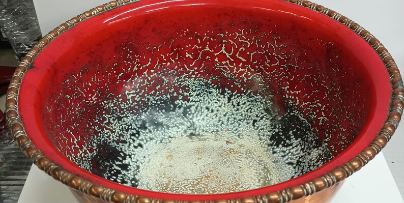 May include: A large, red copper bowl with a white and black crackled glaze. The bowl has a decorative copper rim.