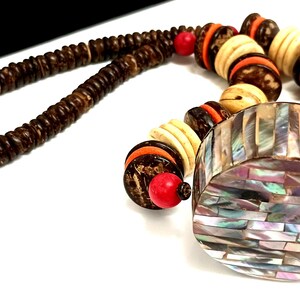 May include: A handcrafted necklace featuring a mix of natural materials. The necklace includes dark brown wooden beads, cream-colored beads with orange accents, and a large, iridescent pendant. Red beads add pops of color.