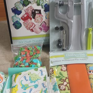 May include: A collection of cloth diaper making supplies, including a book titled "Cloth Diapers Made Easy", snap pliers, colorful snaps, and patterned fabric with dinosaurs and monkeys. The items are arranged on a wooden surface.