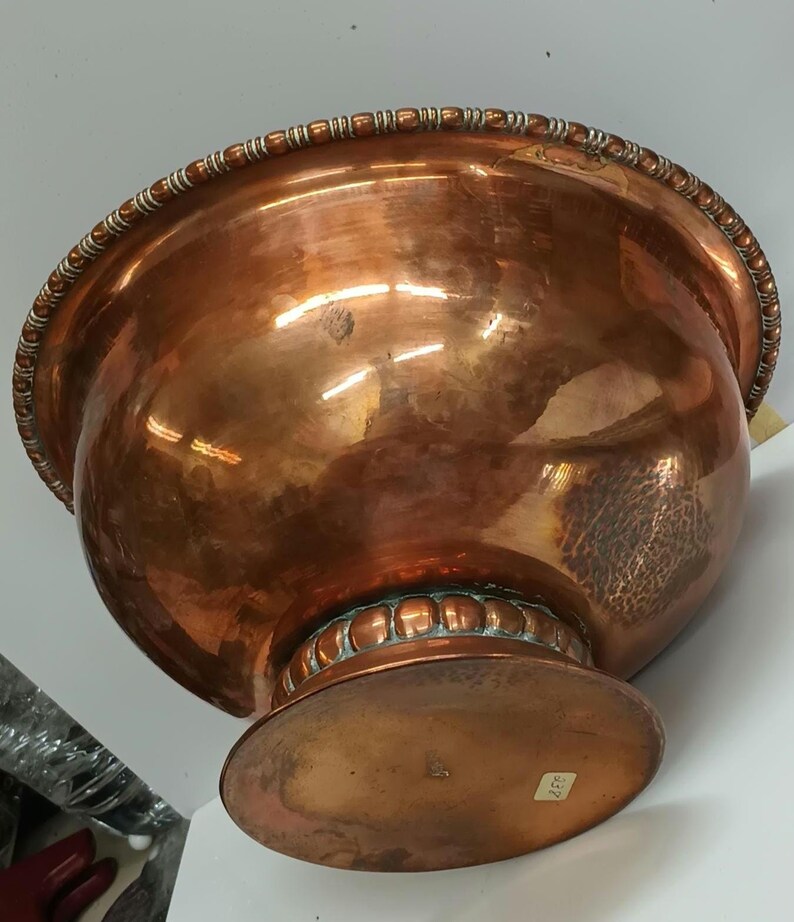 May include: A large copper bowl with a hammered finish and a decorative beaded rim. The bowl sits on a wide, round base.