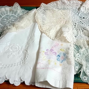 May include: Assortment of vintage white lace and crochet doilies, a small crochet item, and a white embroidered cloth with a floral design. The items are arranged on a wooden surface and a green cutting mat.