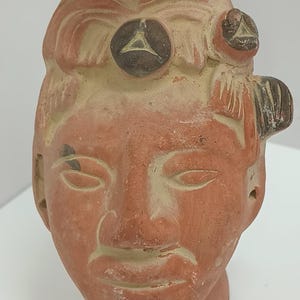 May include: A terracotta sculpture of a human head with a detailed face and hair. The sculpture has two round, dark brown accents on the head, each with a white triangle design.