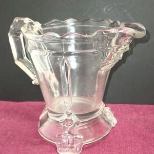 May include: A clear glass creamer with a scalloped rim and a decorative handle. The creamer has a footed base with three decorative feet.