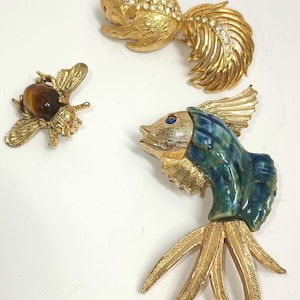 May include: Three gold tone brooches: a bee with a tiger's eye stone, a stylized fish with blue and green enamel, and a stylized fish with rhinestones.