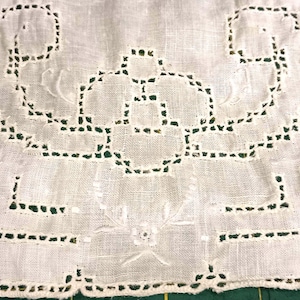 May include: A square white linen napkin featuring intricate cutwork embroidery with a central geometric floral motif and a decorative scalloped border. The fabric has a visible weave texture and the needlework creates openwork patterns throughout.