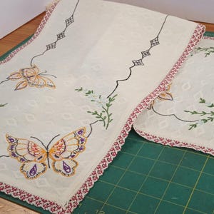 May include: Cream-colored embroidered table runner and matching placemat. The set features butterfly and floral designs in orange, purple, and green. The edges are trimmed with a red and white patterned border.