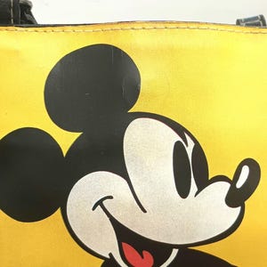 May include: Yellow tote bag featuring a large graphic of Mickey Mouse. The bag has black handles and a black outline around the top. The Mickey Mouse graphic is black, white, and red.