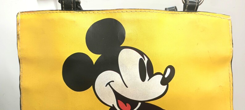 May include: Yellow tote bag featuring a large graphic of Mickey Mouse. The bag has black handles and a black outline around the top. The Mickey Mouse graphic is black, white, and red.