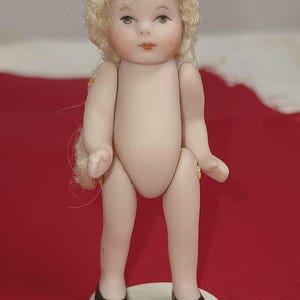 May include: A small doll with a pale, smooth finish and blonde, curly hair. The doll has painted facial features, including dark eyes and red lips. It is wearing black shoes and is standing on a small, circular, off-white base.