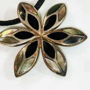 May include: A silver-toned flower pendant with six petals. Each petal features a mother-of-pearl inlay and a black onyx center. The pendant is strung on a black cord, creating a striking contrast.