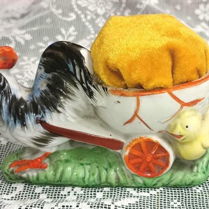 May include: A ceramic figurine featuring a rooster pulling a white cart with orange accents. The cart contains a plush, mustard-yellow velvet pincushion. A small yellow chick is attached to the side of the cart, and the base is painted green.