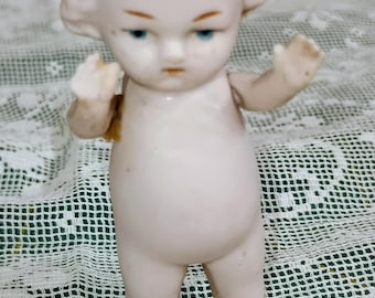 Könnte beinhalten: Eine kleine Figur aus blassem Biskuitporzellan, die ein stehendes Baby mit modelliertem Haar, bemalten blauen Augen und erhobenen Armen darstellt. Die Puppe hat eine glatte, matte Oberfläche und einen runden, pummeligen Körperbau.