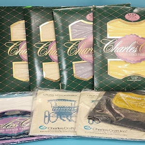 May include: A collection of cross-stitch fabric and supplies. Several green and gold packages of Aida cloth are stacked, with the "Charles Craft" logo. Below are packages of fabric and printed threads, ready for cross-stitch projects.