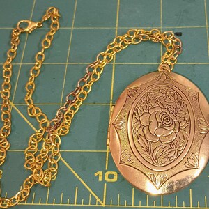 May include: A gold-colored locket necklace with an oval pendant. The pendant features a raised floral design with a rose in the center. The chain is made of linked oval segments and has a clasp.