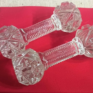 May include: Two clear glass dumbbell-shaped objects with textured handles and faceted ends rest on a red surface. The glass has a diamond-like pattern. The objects appear to be decorative or collectible items.