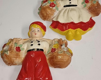 May include: Two ceramic wall hangings depicting figures in traditional Dutch attire, each holding baskets of flowers. The figures wear white tops, red skirts, and yellow shoes. The baskets are brown with colorful flowers.