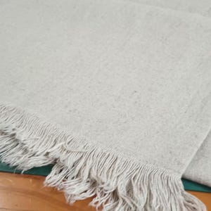 May include: A rectangular piece of light beige, textured fabric with a visible weave and a frayed, fringed edge along one side. The fabric has a natural, unbleached appearance with subtle variations in color throughout the material surface.